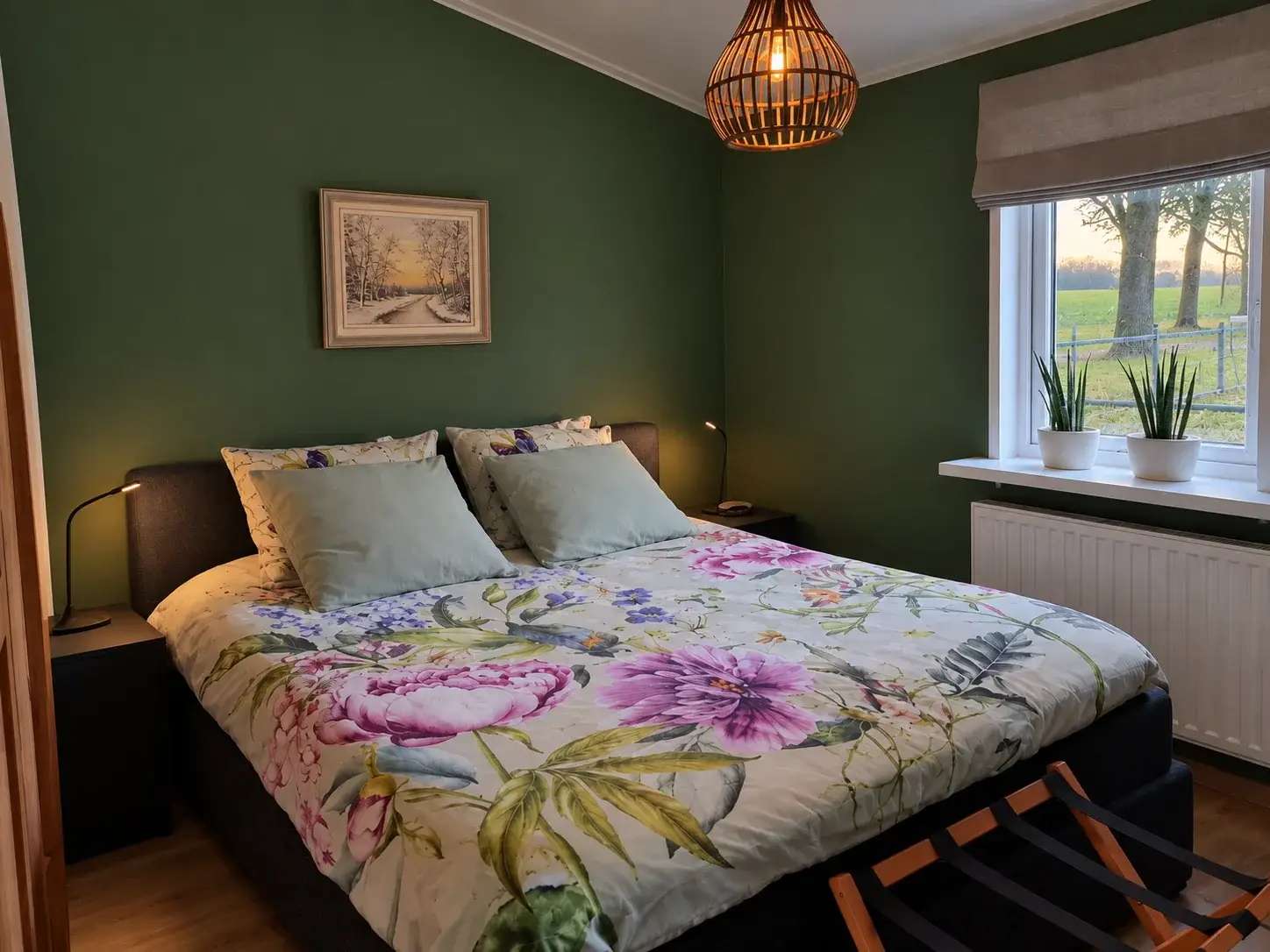 Hoofdslaapkamer met groene wand, tweepersoonsbed met bloemendekbed en raam met uitzicht op de weilanden